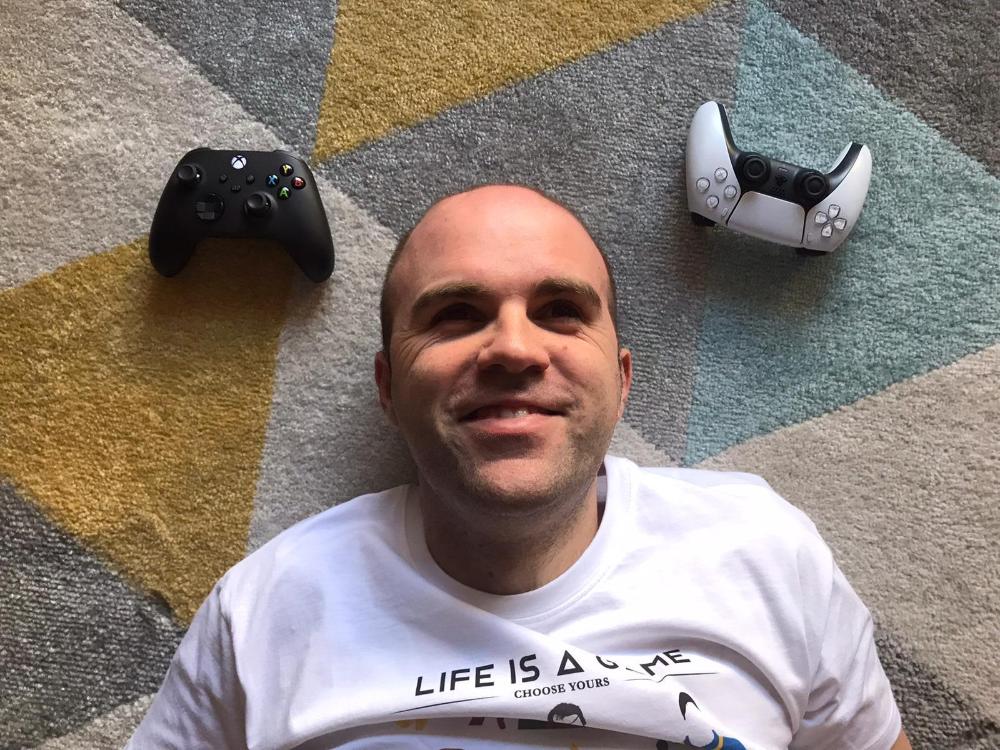 Sergio Vera tumbado en una alfombra con un mando de Xbox a la izquierda y uno de PlayStation a la derecha